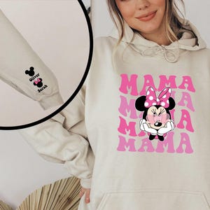 Puede incluir: Una sudadera con capucha beige con un gr&aacute;fico rosa y blanco de Mickey Mouse con el texto "MAMA" repetido cuatro veces. La sudadera tiene un bolsillo en la parte delantera y una cabeza de Mickey Mouse negra con el texto "Oliver" y "Sarah" en la manga.