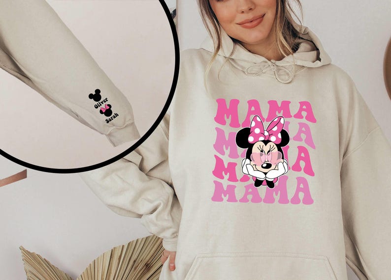 Puede incluir: Una sudadera con capucha beige con un gr&aacute;fico rosa y blanco de Mickey Mouse con el texto "MAMA" repetido cuatro veces. La sudadera tiene un bolsillo en la parte delantera y una cabeza de Mickey Mouse negra con el texto "Oliver" y "Sarah" en la manga.