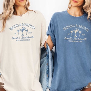 May include: Two t-shirts, one ivory and one blue jean, with the text "Bikinis & Martinis est 2024 Sarah's Bachelorette Tulum Beach Club" printed on the front.
