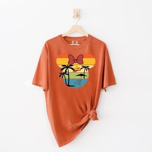 May include: A burnt orange t-shirt with a retro sunset graphic featuring a palm tree and a red bow. The graphic is in the shape of a mouse head.