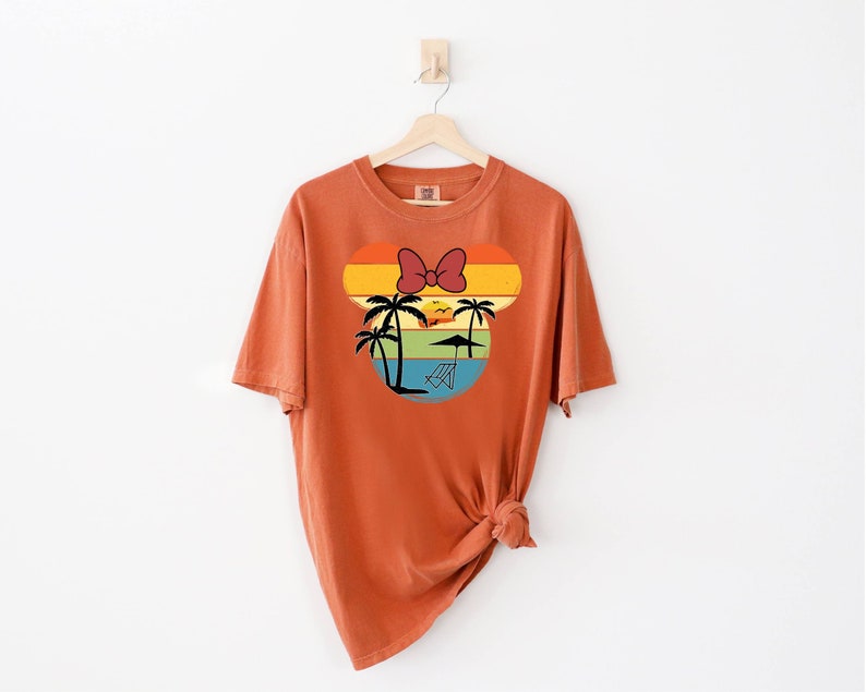 May include: A burnt orange t-shirt with a retro sunset graphic featuring a palm tree and a red bow. The graphic is in the shape of a mouse head.