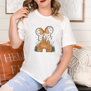 Puede incluir: Camiseta blanca con un dise&ntilde;o de castillo de jengibre. El castillo est&aacute; decorado con un lazo rojo, estrellas y fuegos artificiales. El dise&ntilde;o recuerda a un parque de atracciones popular.