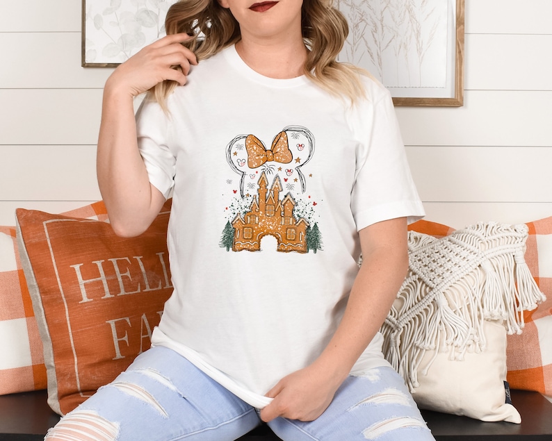 Puede incluir: Camiseta blanca con un dise&ntilde;o de castillo de jengibre. El castillo est&aacute; decorado con un lazo rojo, estrellas y fuegos artificiales. El dise&ntilde;o recuerda a un parque de atracciones popular.
