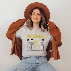 May include: A light gray t-shirt with the word "ADHD" in yellow, along with three skeleton figures. Two skeletons are standing, and one is dabbing. The text "Why fit in when you were born to stand out!" is below the skeletons.