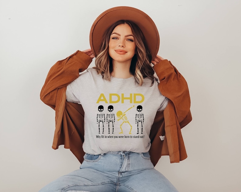 May include: A light gray t-shirt with the word "ADHD" in yellow, along with three skeleton figures. Two skeletons are standing, and one is dabbing. The text "Why fit in when you were born to stand out!" is below the skeletons.