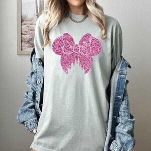 May include: A light green t-shirt with a pink glitter bow design. The bow is shaped like Mickey Mouse ears.