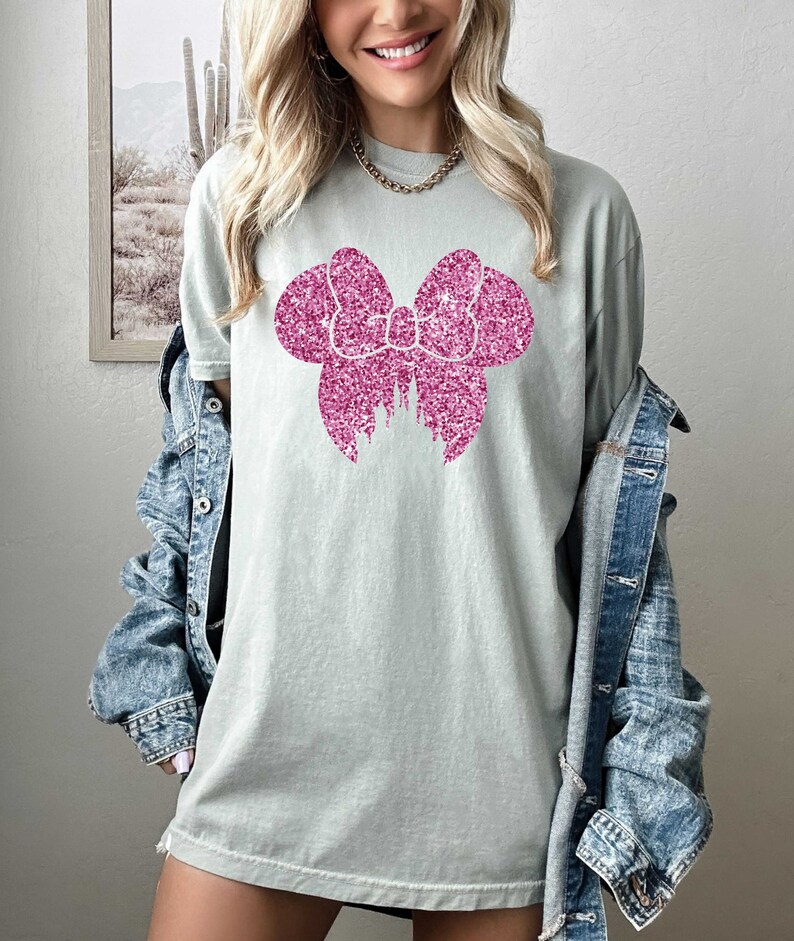 May include: A light green t-shirt with a pink glitter bow design. The bow is shaped like Mickey Mouse ears.