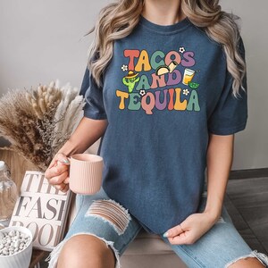 Puede incluir: Una camiseta azul marino con el texto "Tacos and Tequila" en colores arco&iacute;ris. El texto est&aacute; en un estilo de fuente retro con un gr&aacute;fico de cactus y taco.