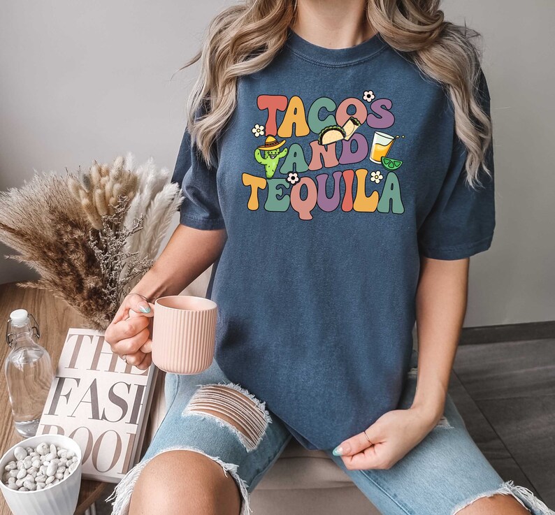 Puede incluir: Una camiseta azul marino con el texto "Tacos and Tequila" en colores arco&iacute;ris. El texto est&aacute; en un estilo de fuente retro con un gr&aacute;fico de cactus y taco.