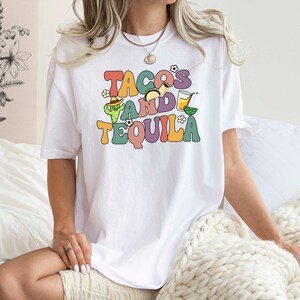 Puede incluir: Camiseta blanca con un gr&aacute;fico colorido que dice "Tacos and Tequila" con ilustraciones de un taco, una margarita y un cactus.