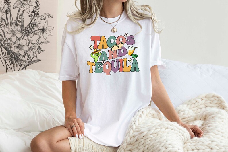 Puede incluir: Camiseta blanca con un gr&aacute;fico colorido que dice "Tacos and Tequila" con ilustraciones de un taco, una margarita y un cactus.