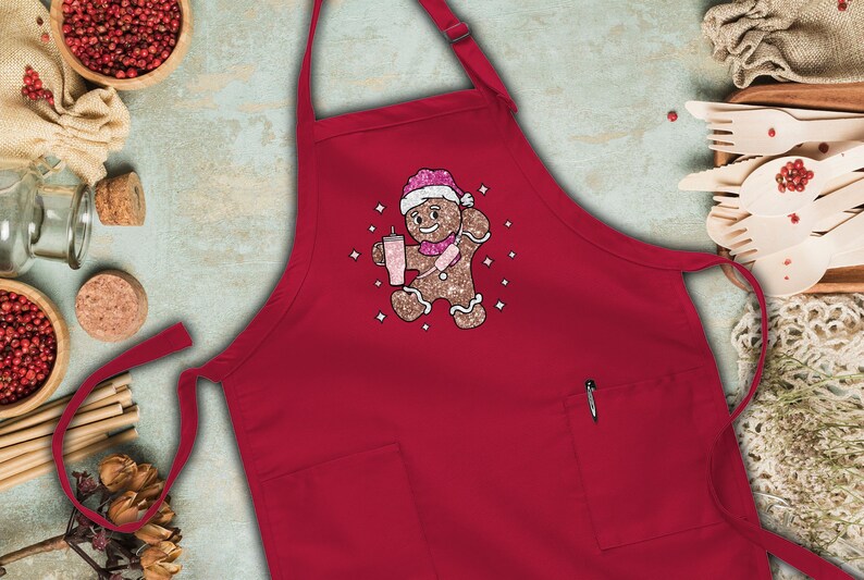 Peut inclure: Tablier de cuisine rouge avec un bonhomme en pain d'&eacute;pices portant un chapeau de P&egrave;re No&euml;l et tenant une boisson rose. Le tablier a une poche sur le c&ocirc;t&eacute; droit.