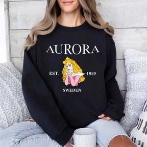 Peut inclure: Sweat-shirt noir avec le mot "AURORA" en blanc, une illustration de princesse et le texte "EST. 1959 SWEDEN". Le sweat-shirt est de style col rond. La personne porte le sweat-shirt.