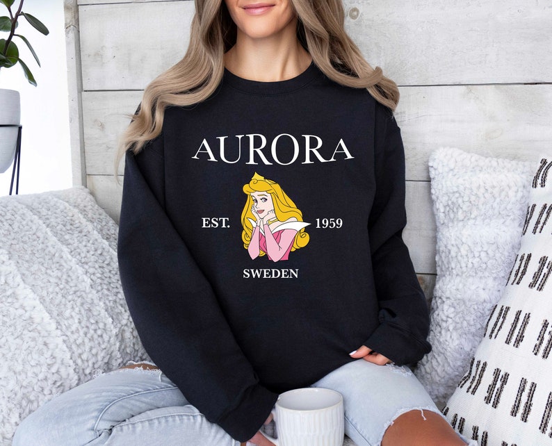 Peut inclure: Sweat-shirt noir avec le mot "AURORA" en blanc, une illustration de princesse et le texte "EST. 1959 SWEDEN". Le sweat-shirt est de style col rond. La personne porte le sweat-shirt.