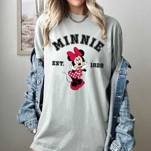 May include: A light green t-shirt with a black and white graphic of Minnie Mouse. The graphic includes the text "MINNIE EST. 1928".