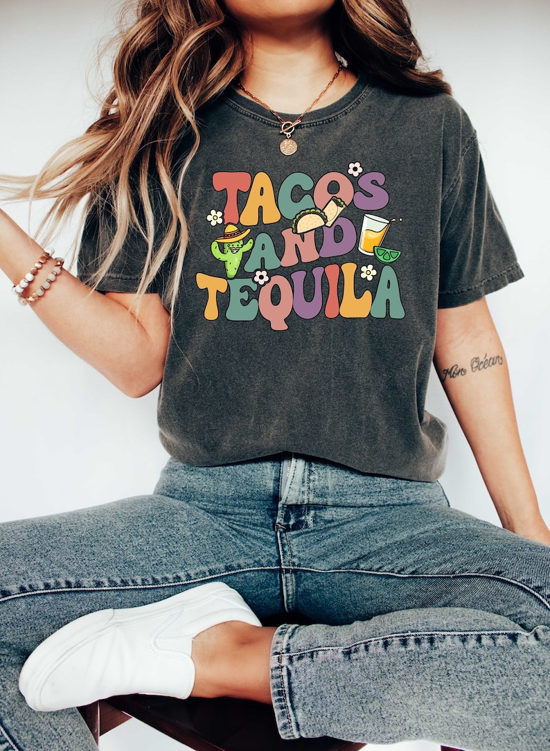 Puede incluir: Una camiseta gris oscuro con un gr&aacute;fico colorido que dice "Tacos and Tequila" con un cactus, tacos y una copa de margarita.