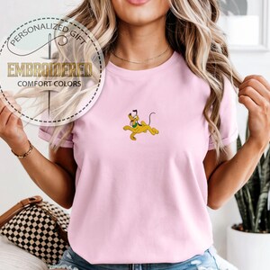 May include: A light pink t-shirt with an embroidered Pluto graphic. The shirt has short sleeves and a crew neck. The text "PERSONALIZED GIFT EMBROIDERED COMFORT COLORS" is printed on a circular label.
