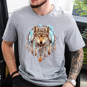 Pode incluir: Camiseta cinza com um gr&aacute;fico de um lobo usando uma coroa de apanhadores de sonhos. O lobo &eacute; marrom e branco com olhos azuis.