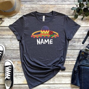 K&ouml;nnte beinhalten: Ein dunkelgraues T-Shirt mit einem bunten Sombrero und einer roten Rosengrafik. Der Text "NAME" ist unter dem Sombrero gedruckt.