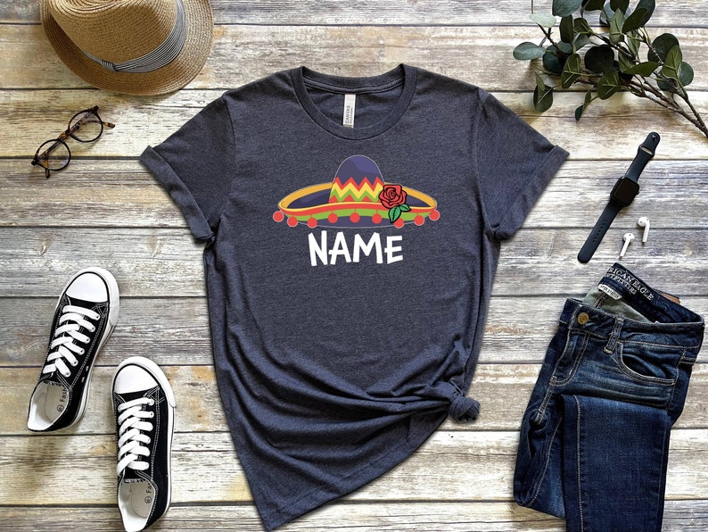 K&ouml;nnte beinhalten: Ein dunkelgraues T-Shirt mit einem bunten Sombrero und einer roten Rosengrafik. Der Text "NAME" ist unter dem Sombrero gedruckt.