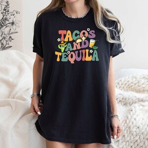 Puede incluir: Camiseta negra con un gr&aacute;fico colorido que dice "Tacos and Tequila" con un cactus, tacos y una copa de margarita.