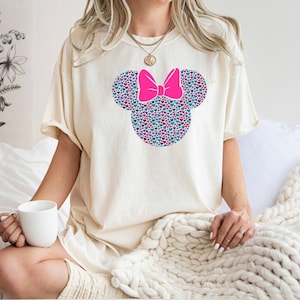 以下が含まれることがあります： カラフルなハート型のミニーマウスのデザインが施された白いTシャツ。デザインにはピンクのリボンが付いており、シャツの中央に配置されています。