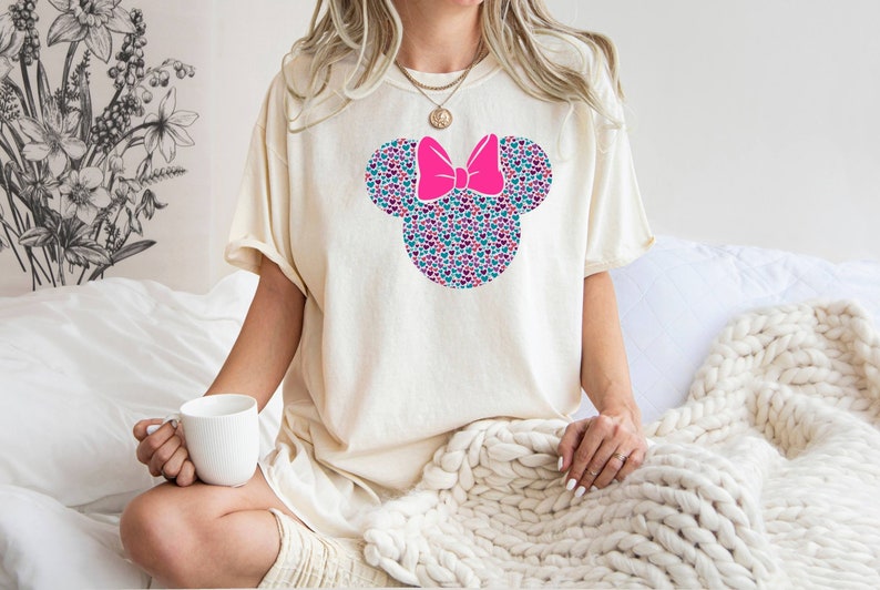 以下が含まれることがあります： カラフルなハート型のミニーマウスのデザインが施された白いTシャツ。デザインにはピンクのリボンが付いており、シャツの中央に配置されています。