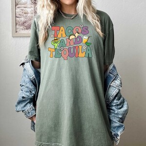 Puede incluir: Una camiseta verde con un gr&aacute;fico de estilo retro que dice "Tacos and Tequila" con ilustraciones de tacos, un cactus, un sombrero y una copa de margarita.
