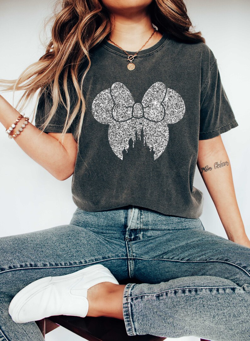 May include: A charcoal grey t-shirt with a silver glitter bow and a castle silhouette. The bow is in the shape of Minnie Mouse's ears.