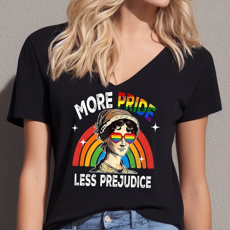 May include: Black v-neck t-shirt with a rainbow and a woman's face wearing rainbow heart-shaped sunglasses. The text "More Pride Less Prejudice" is printed on the shirt.