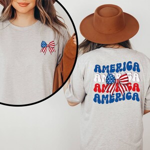 K&ouml;nnte beinhalten: Ein graues T-Shirt mit einer rot-wei&szlig;-blauen Schleife mit Sternen auf der Vorderseite und dem Wort "AMERICA" viermal in einer wellenf&ouml;rmigen Schrift auf der R&uuml;ckseite. Das Wort "AMERICA" ist in Blau, Wei&szlig; und Rot.