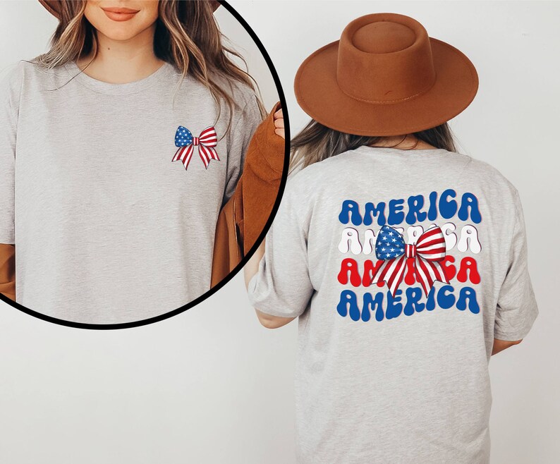 K&ouml;nnte beinhalten: Ein graues T-Shirt mit einer rot-wei&szlig;-blauen Schleife mit Sternen auf der Vorderseite und dem Wort "AMERICA" viermal in einer wellenf&ouml;rmigen Schrift auf der R&uuml;ckseite. Das Wort "AMERICA" ist in Blau, Wei&szlig; und Rot.