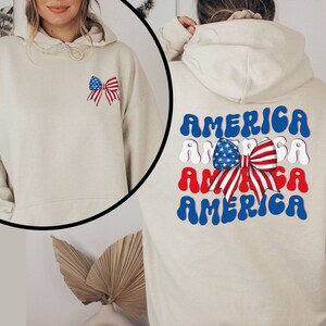 K&ouml;nnte beinhalten: Ein beiger Hoodie mit einer rot-wei&szlig;-blauen Schleife mit Sternen auf der linken Brust und dem Wort "AMERICA", das viermal in einer wellenf&ouml;rmigen Schrift auf dem R&uuml;cken wiederholt wird.