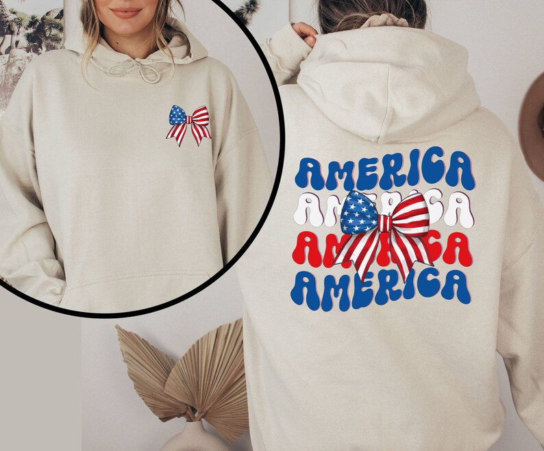 K&ouml;nnte beinhalten: Ein beiger Hoodie mit einer rot-wei&szlig;-blauen Schleife mit Sternen auf der linken Brust und dem Wort "AMERICA", das viermal in einer wellenf&ouml;rmigen Schrift auf dem R&uuml;cken wiederholt wird.