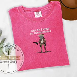 May include: A hot pink Comfort Colors t-shirt with the embroidered phrase "Hold On Partner I'm Overstimulated" and a cartoon frog wearing a cowboy hat and holding a gun. The shirt is a personalized gift.