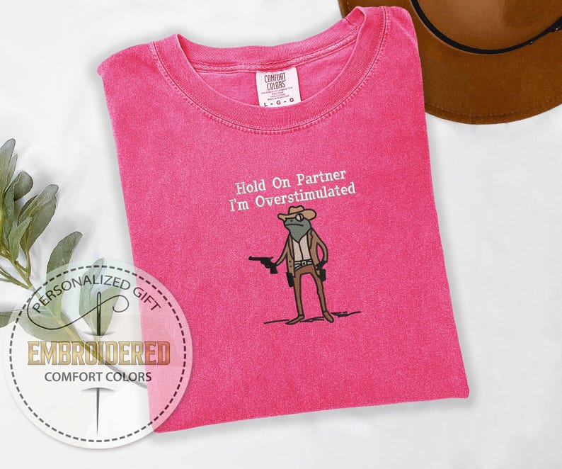 May include: A hot pink Comfort Colors t-shirt with the embroidered phrase "Hold On Partner I'm Overstimulated" and a cartoon frog wearing a cowboy hat and holding a gun. The shirt is a personalized gift.