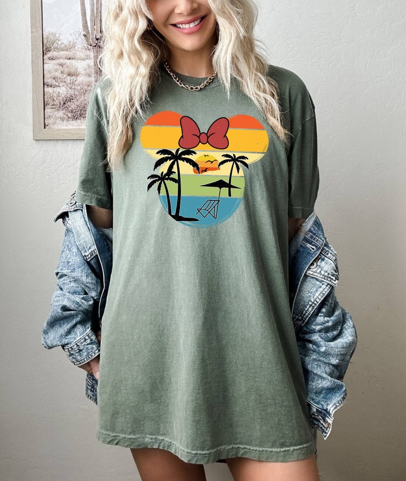 May include: A green t-shirt with a retro-style graphic of a cartoon mouse head with a red bow, palm trees, a sunset, and a beach chair. The graphic is in shades of orange, yellow, blue, and green.