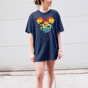 May include: A navy blue t-shirt with a vintage-style graphic of a Mickey Mouse head silhouette. The graphic features a sunset with palm trees and a beach umbrella.
