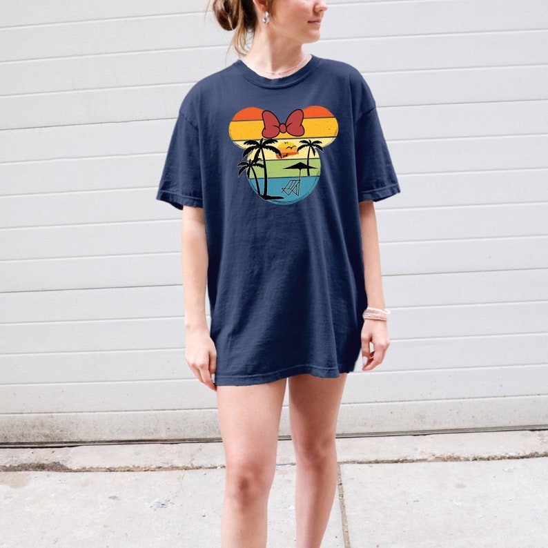 May include: A navy blue t-shirt with a vintage-style graphic of a Mickey Mouse head silhouette. The graphic features a sunset with palm trees and a beach umbrella.