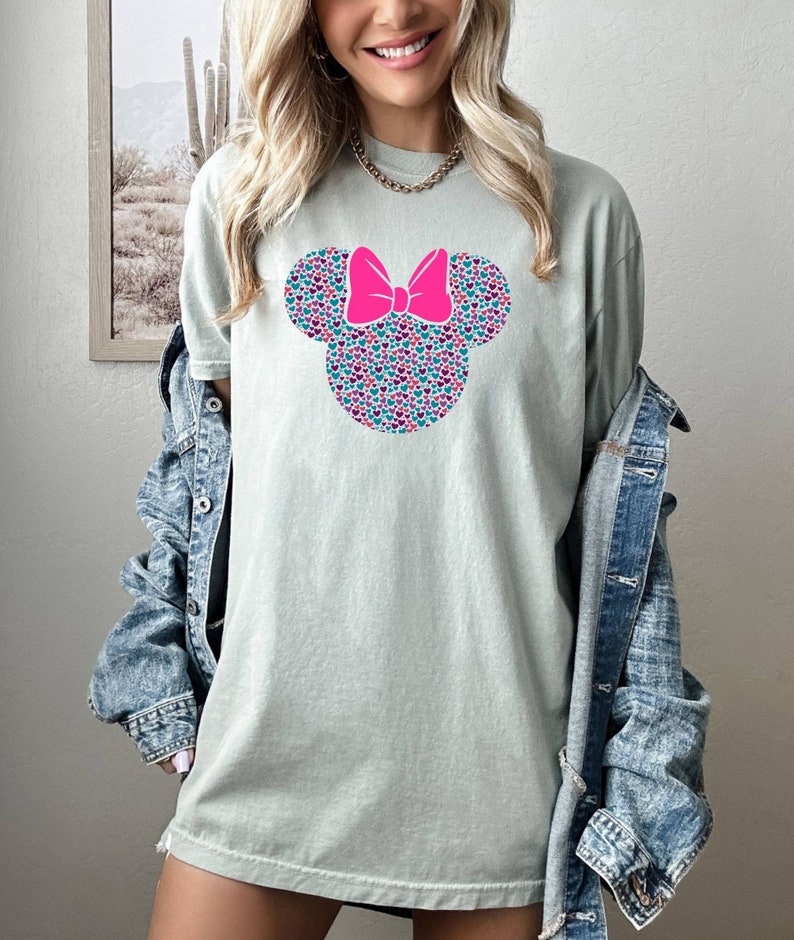 以下が含まれることがあります： 薄い緑色のTシャツに、ピンクのリボンと、ピンク、青、紫の小さなハートでできたミッキーマウスの頭のグラフィックが描かれています。