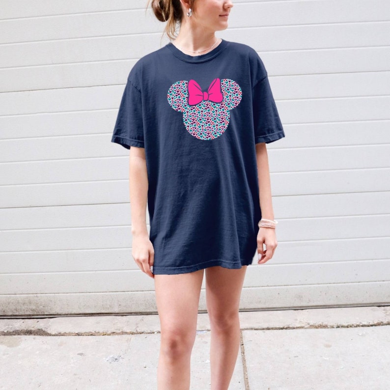 以下が含まれることがあります： カラフルなミニーマウスの頭のグラフィックがプリントされたネイビーブルーのTシャツ。グラフィックは、小さくカラフルなドットで構成されており、ピンクのリボンが付いています。