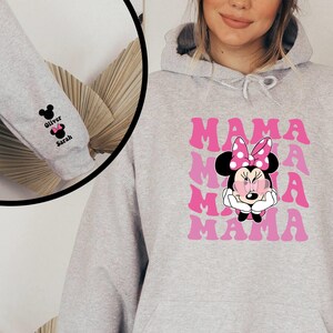 Puede incluir: Sudadera gris con un gr&aacute;fico rosa y blanco de Minnie Mouse y la palabra "MAMA" repetida. La manga tiene un gr&aacute;fico en blanco y negro de Minnie Mouse con los nombres "Oliver" y "Sarah" debajo.