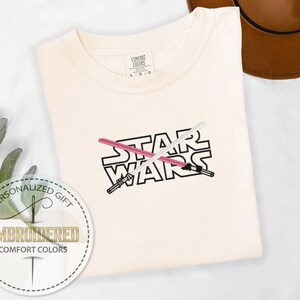 May include: A white t-shirt with the words "STAR WARS" embroidered in black with two crossed lightsabers in pink and white.