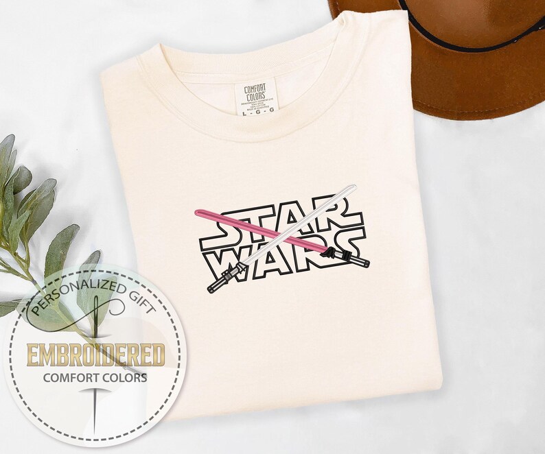 May include: A white t-shirt with the words "STAR WARS" embroidered in black with two crossed lightsabers in pink and white.
