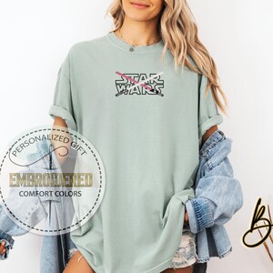 May include: A light green t-shirt featuring the text "STAR WARS" and a pink and white lightsaber design. The t-shirt is embroidered with the text "PERSONALIZED GIFT EMBROIDERED COMFORT COLORS".