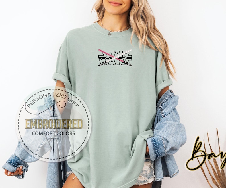 May include: A light green t-shirt featuring the text "STAR WARS" and a pink and white lightsaber design. The t-shirt is embroidered with the text "PERSONALIZED GIFT EMBROIDERED COMFORT COLORS".