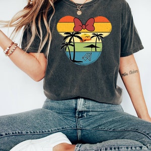 May include: A gray t-shirt with a vintage-style graphic of Mickey Mouse's head. The graphic features a sunset, palm trees, and a beach chair. The shirt has a red bow on top of the head.