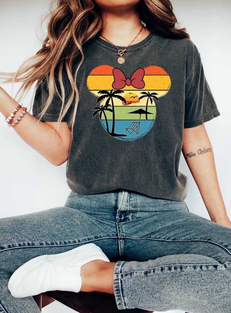 May include: A gray t-shirt with a vintage-style graphic of Mickey Mouse's head. The graphic features a sunset, palm trees, and a beach chair. The shirt has a red bow on top of the head.
