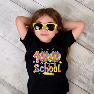 May include: A black t-shirt with a colorful graphic that says "100 day of school" with a pencil and apples. A child is wearing the shirt and has yellow sunglasses on.