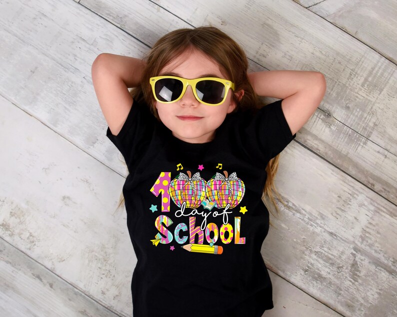 May include: A black t-shirt with a colorful graphic that says "100 day of school" with a pencil and apples. A child is wearing the shirt and has yellow sunglasses on.